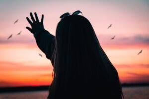 Silhouette of a woman reaching towards seagulls against a vibrant sunset sky.