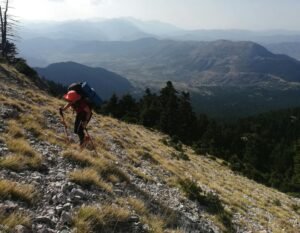A hiker climbs a rocky mountain trail under bright daylight, with expansive scenic views.
